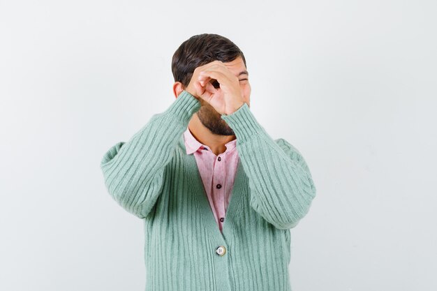 Junger Mann, der vorgibt, mit seinen Händen im Hemd, in der Strickjacke durch ein Loch zu spähen und fokussiert zu schauen, Vorderansicht.