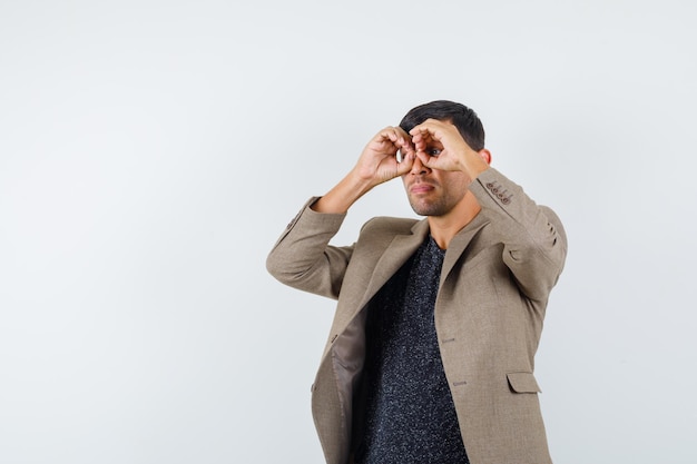 Junger Mann, der wegschaut, während er eine Brille zeigt, die mit seinen Fingern in graubrauner Jacke, schwarzem Hemd und seltsam aussieht.