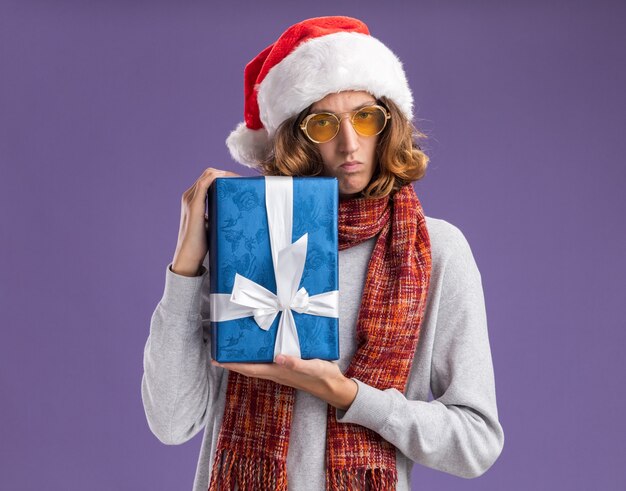 Junger Mann, der Weihnachtsweihnachtsmütze und gelbe Brille mit warmem Schal um seinen Hals hält, der Weihnachtsgeschenk hält und Kamera mit ernstem Gesicht über lila Hintergrund steht