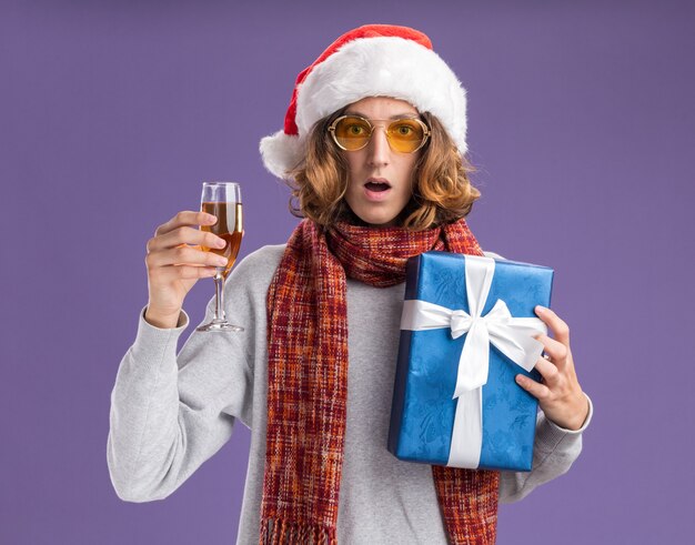 junger Mann, der Weihnachtsweihnachtsmütze und gelbe Brille mit warmem Schal um seinen Hals hält, der Weihnachtsgeschenk und Glas Champagner hält Kamera betrachtet überrascht über lila Hintergrund steht