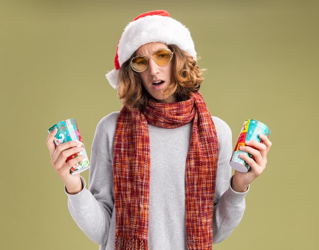 junger Mann, der Weihnachtsweihnachtsmütze und gelbe Brille mit warmem Schal um seinen Hals trägt, der bunte Pappbecher hält, die verwirrt schauen und Zweifel haben, die über grünem Hintergrund stehen