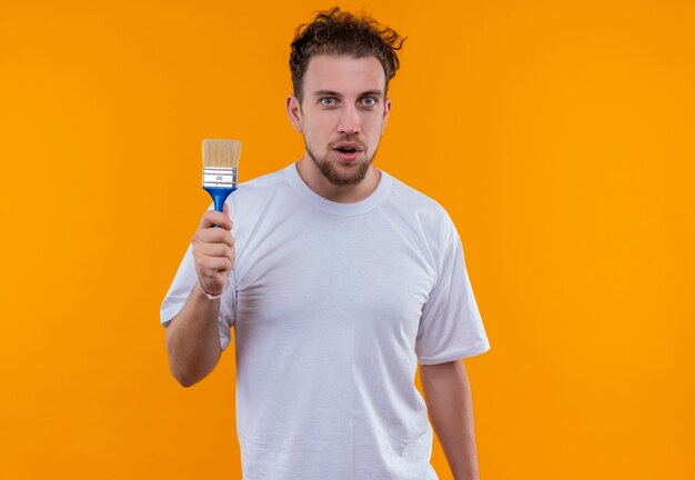 junger Mann, der weißen T-Shirt hält Pinsel auf isolierter orange Wand hält