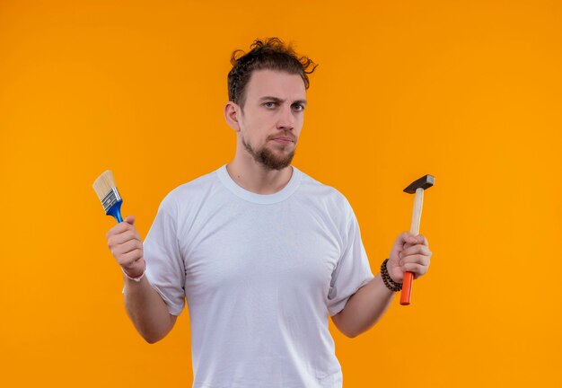 junger Mann, der weißes T-Shirt hält, das Pinsel und Hammer auf isolierter orange Wand hält