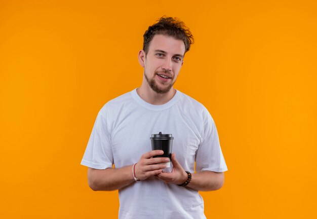 junger Mann, der weißes T-Shirt hält, das Tasse Kaffee auf isolierter orange Wand hält