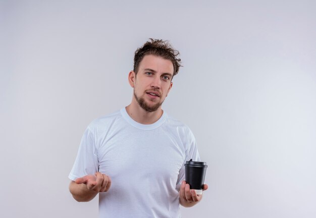 junger Mann, der weißes T-Shirt hält, das Tasse Kaffee hält, der Sie Geste auf isolierter weißer Wand zeigt