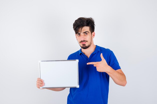 Junger Mann, der Whiteboard hält und darauf in blauem T-Shirt zeigt und ernst aussieht
