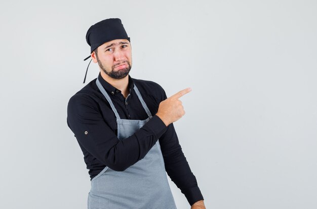 Junger Mann, der zur Seite in Hemd, Schürze zeigt und beleidigt schaut, Vorderansicht.