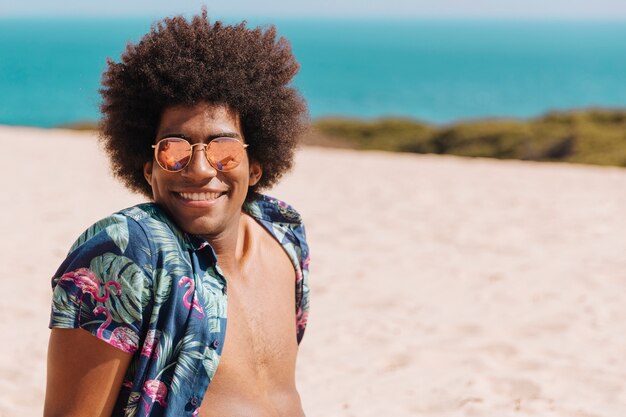 Junger Mann des Afroamerikaners in der Sonnenbrille, die Kamera auf Strand betrachtet