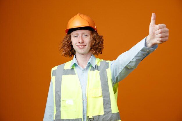 Junger Mann des Baumeisters in Bauuniform und Schutzhelm, der glücklich und selbstbewusst lächelnd in die Kamera schaut und den Daumen nach oben zeigt, der über orangefarbenem Hintergrund steht