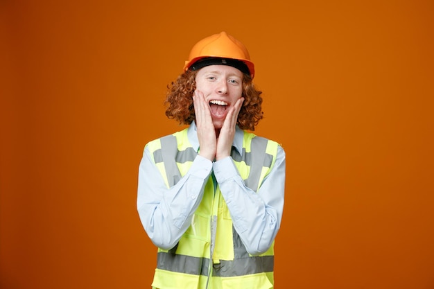 Junger Mann des Baumeisters in Bauuniform und Schutzhelm, der glücklich und zufrieden lächelnd in die Kamera schaut und breit die Hände auf seinen Wangen hält, die über orangefarbenem Hintergrund stehen