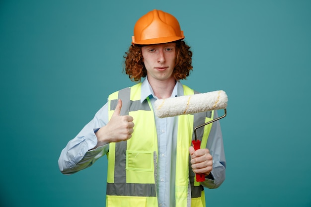 Junger Mann des Baumeisters in Bauuniform und Schutzhelm mit Farbroller, der in die Kamera blickt und selbstbewusst lächelt und Daumen nach oben zeigt, der über blauem Hintergrund steht