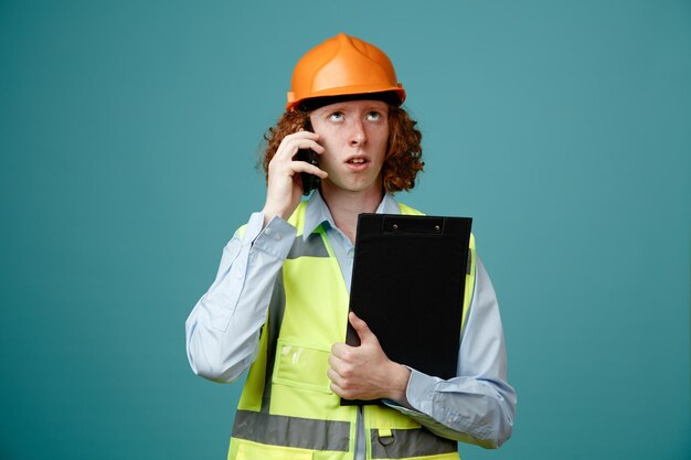 Junger Mann des Baumeisters in Bauuniform und Schutzhelm mit Klemmbrett, der am Handy spricht und verwirrt über blauem Hintergrund steht