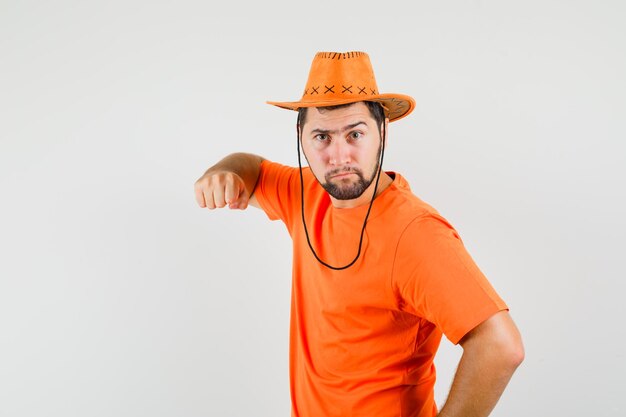 Junger Mann droht mit Faust in orangefarbenem T-Shirt, Hut und sieht wütend aus, Vorderansicht.
