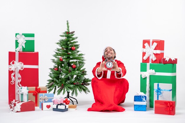 Junger Mann feiern Weihnachtsfeiertag, der im Boden sitzt und Uhr nahe Geschenken und geschmücktem Weihnachtsbaum zeigt