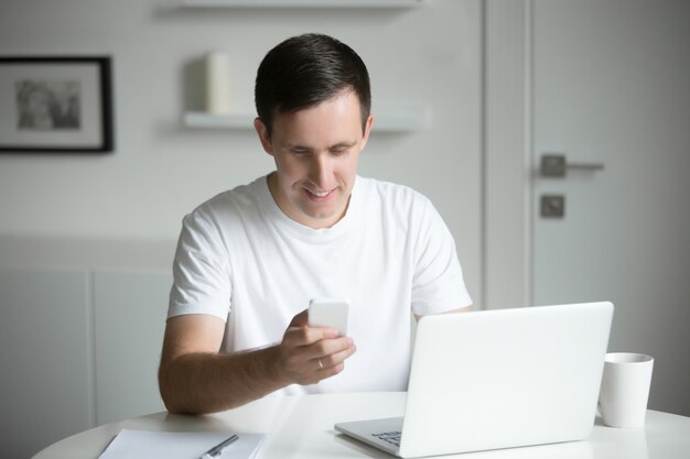 Junger Mann hält ein Telefon, sitzt am Schreibtisch, Laptop in der Nähe