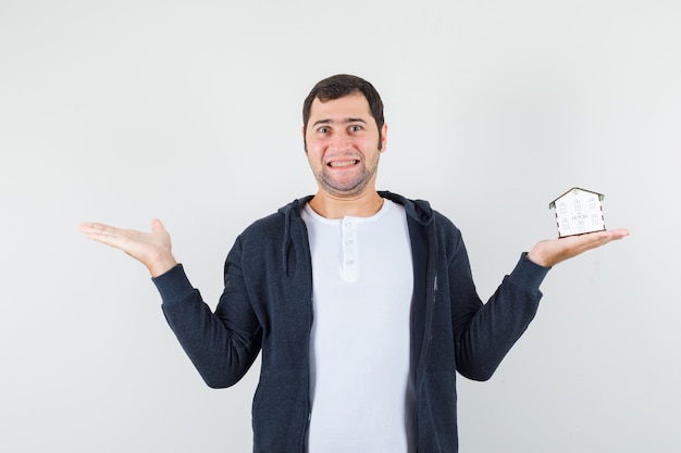 Junger Mann hält Hausmodell in einer Hand und streckt eine andere Hand nach links in weißem T-Shirt und schwarzem Kapuzenpullover mit Reißverschluss vorne und sieht optimistisch aus, Vorderansicht.