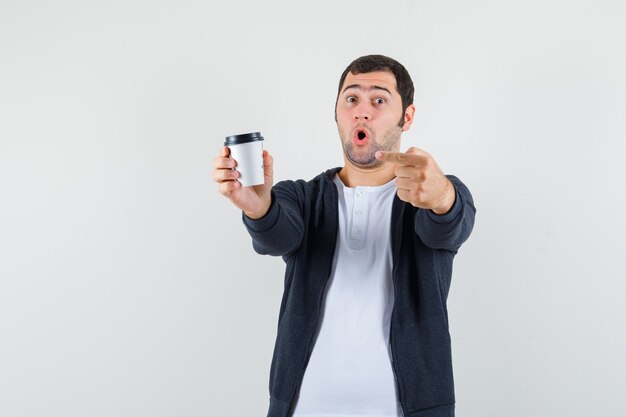 Junger Mann hält Kaffeetasse zum Mitnehmen und zeigt mit dem Zeigefinger auf das weiße T-Shirt und den schwarzen Kapuzenpulli mit Reißverschluss und sieht überrascht aus, Vorderansicht.