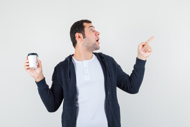 Junger Mann hält Kaffeetasse zum Mitnehmen und zeigt nach rechts mit dem Zeigefinger in weißem T-Shirt und schwarzem Kapuzenpullover mit Reißverschluss vorne und sieht überrascht aus, Vorderansicht.