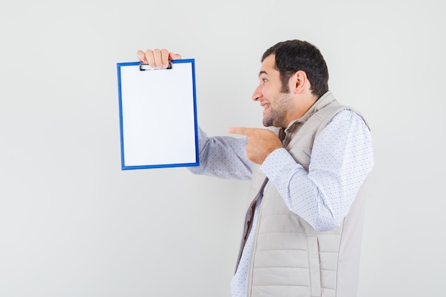 Junger Mann hält Notizbuch und zeigt darauf in beige Jacke und schaut optimistisch, Vorderansicht.