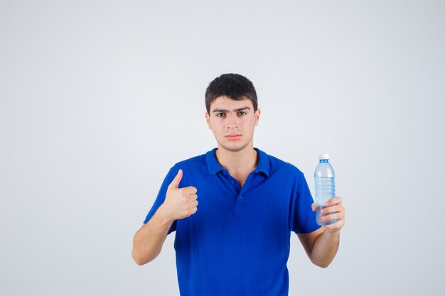 Junger Mann hält Plastikflasche, zeigt Daumen oben im T-Shirt und schaut selbstbewusst, Vorderansicht.