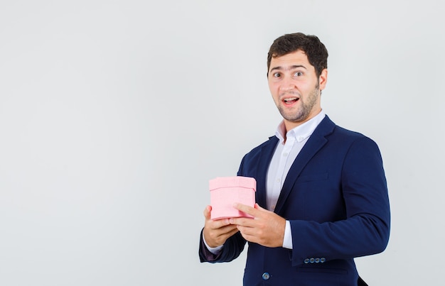 Junger Mann hält rosa Geschenkbox im Anzug und schaut fröhlich, Vorderansicht.