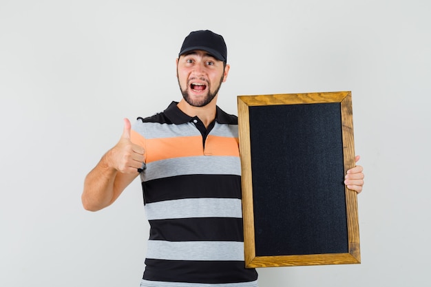 Kostenloses Foto junger mann hält tafel, zeigt daumen oben in t-shirt, mütze und schaut glücklich. vorderansicht.