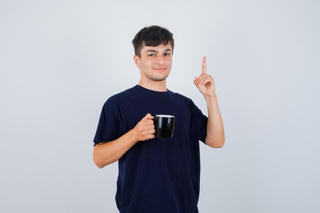 Junger Mann hält Tasse Tee, zeigt in schwarzem T-Shirt und sieht selbstbewusst aus. Vorderansicht.