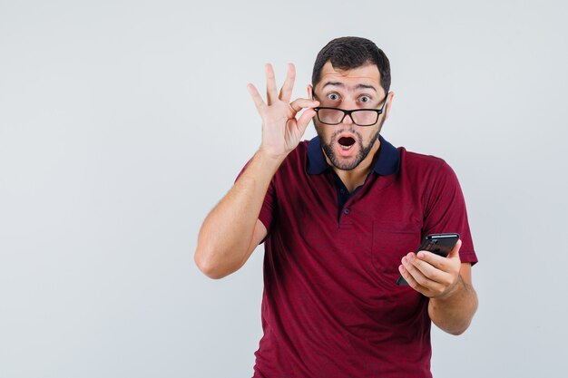 Junger Mann hält Telefon im roten T-Shirt, Brille und schaut erstaunt, Vorderansicht.