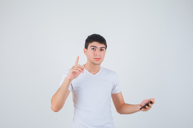 Junger Mann hält Telefon, zeigt in T-Shirt und schaut selbstbewusst, Vorderansicht.