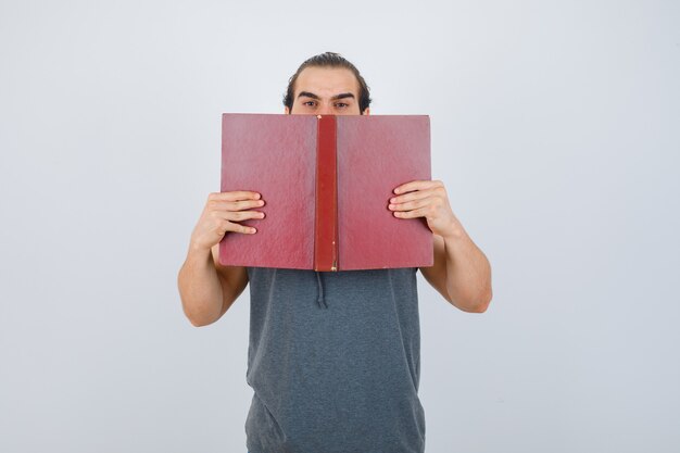 Junger Mann im ärmellosen Kapuzenpulli, der geöffnetes Buch auf Mund hält und ernsthafte Vorderansicht sieht.