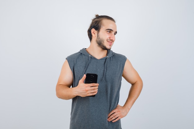 Junger Mann im ärmellosen Kapuzenpulli, der Handy hält, während Hand auf Hüfte hält und gut aussehend, Vorderansicht schaut.