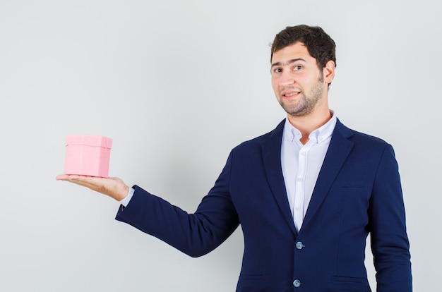 Junger Mann im Anzug hält Geschenkbox und schaut fröhlich, Vorderansicht.