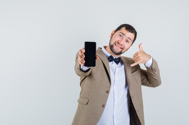 Junger Mann im Anzug zeigt Handy mit Anrufgeste und schaut fröhlich, Vorderansicht.