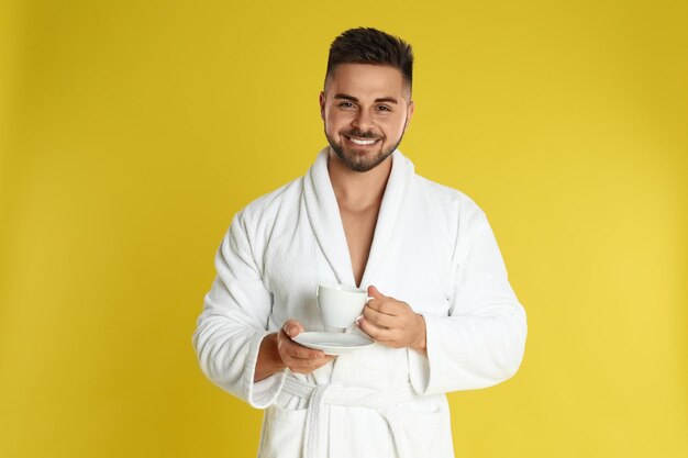 Junger Mann im Bademantel mit Tasse Kaffee auf gelbem Hintergrund