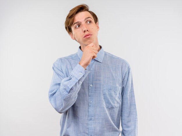 Junger Mann im blauen Hemd, das beiseite mit Hand auf Kinn denkt, das über weißer Wand steht
