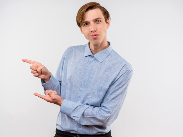 Junger Mann im blauen Hemd, das mit Zeigefingern auf die Seite zeigt, die zuversichtlich weiß über der weißen Wand steht