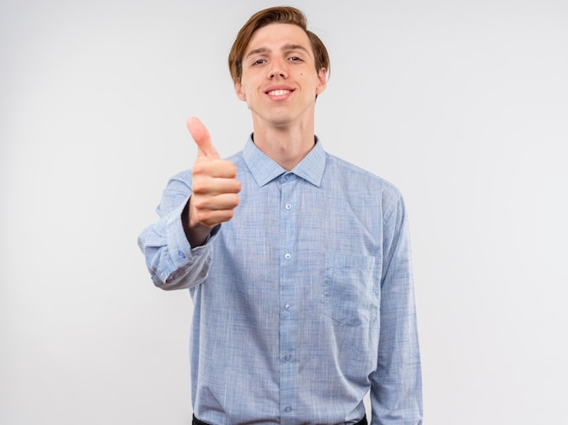 Junger Mann im blauen Hemd lächelnd mit glücklichem afce, das Daumen oben steht über weißer Wand zeigt
