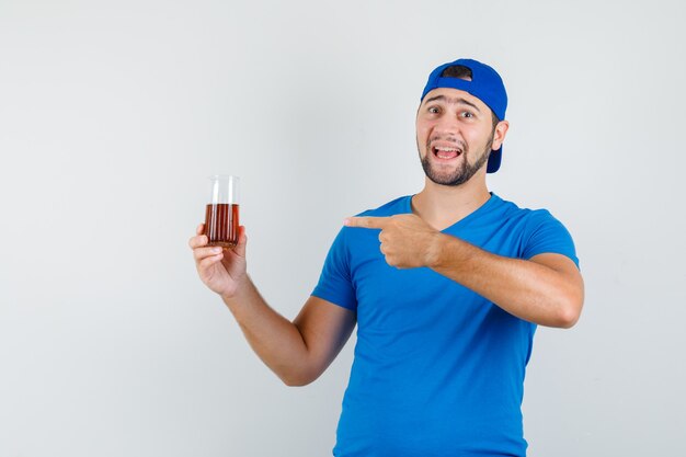 Junger Mann im blauen T-Shirt und in der Kappe, die auf Glas des Getränks zeigen