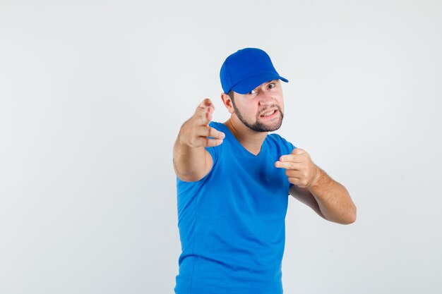 Junger Mann im blauen T-Shirt und in der Kappe, die durch Waffengeste auf Kamera zeigen und wütend schauen