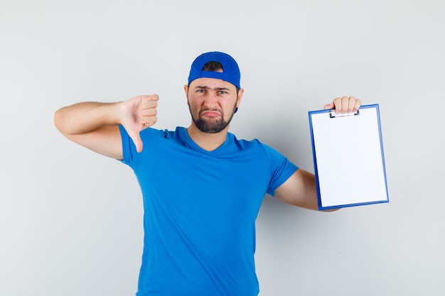 Junger Mann im blauen T-Shirt und in der Kappe, die Klemmbrett mit Daumen nach unten halten und unzufrieden aussehen