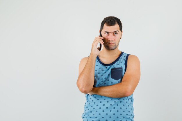 Junger Mann im blauen Unterhemd, der auf Handy spricht und zögernd schaut