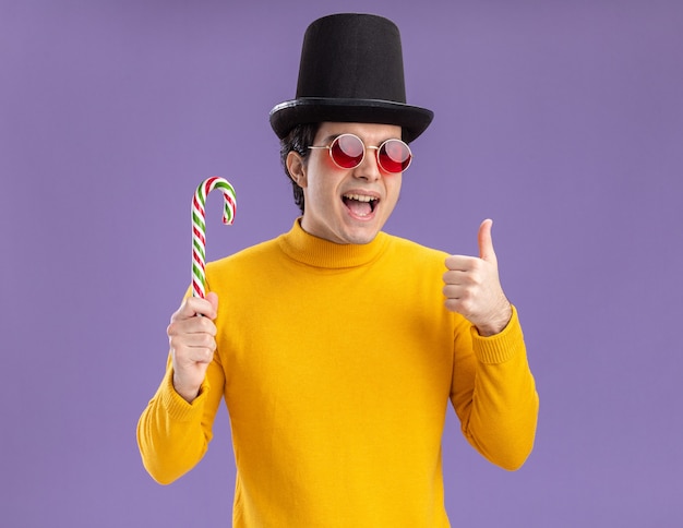 Junger Mann im gelben Rollkragenpullover und in den Gläsern, die schwarzen Hut tragen, der Zuckerstange hält, der fröhlich zeigt Daumen zeigt, die über lila Wand stehen