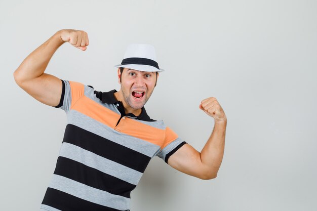 Junger Mann im gestreiften T-Shirt, Hut hebt die Hände, während er seine Kraft zeigt und energisch aussieht