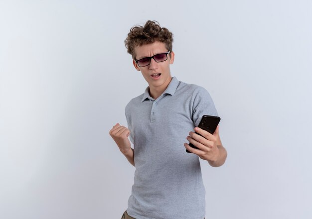 Junger Mann im grauen Poloshirt, der seinen Smartphonebildschirm betrachtet, der die Faust ballt, die seinen Erfolg erfreut, der über weißer Wand steht