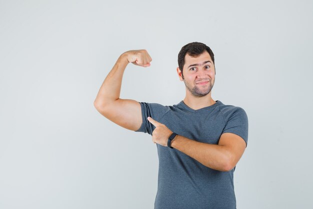 Junger Mann im grauen T-Shirt, der auf Armmuskeln zeigt und zuversichtlich schaut