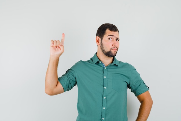 Kostenloses Foto junger mann im grünen hemd, der mit fragender art nach oben zeigt und fokussiert aussieht, vorderansicht.