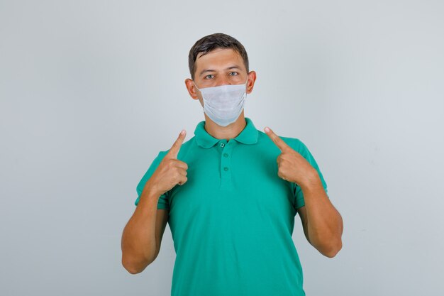 Junger Mann im grünen T-Shirt zeigt seine medizinische Maske und schaut vorsichtig, Vorderansicht.