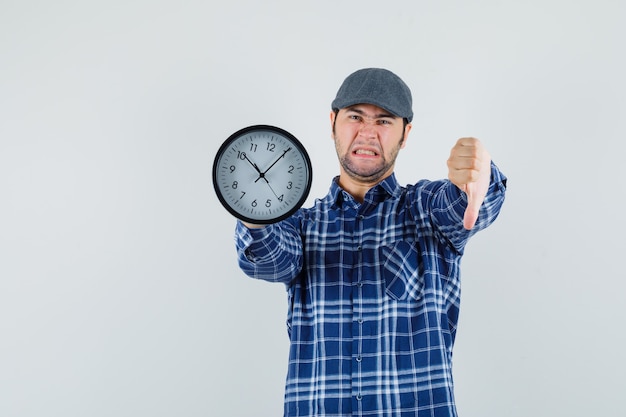 Junger Mann im Hemd, Kappe, die Wanduhr hält, Daumen nach unten zeigt und Unzufriedenheit schaut, Vorderansicht.