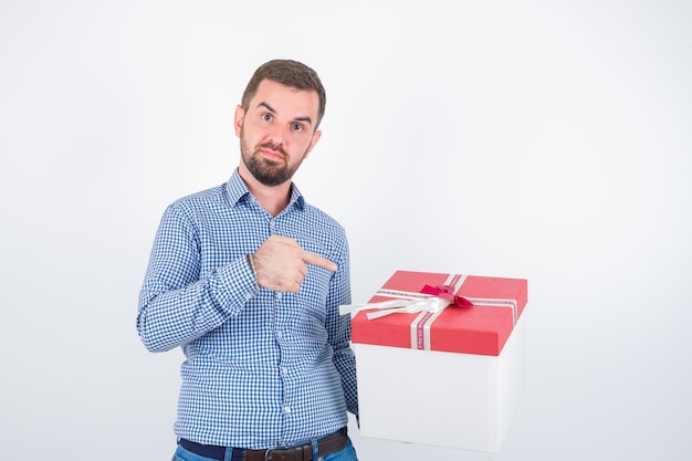 Junger Mann im Hemd zeigt auf Geschenkbox und schaut selbstbewusst, Vorderansicht.