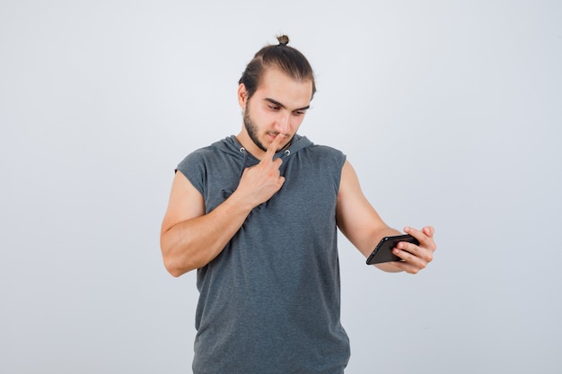 Junger Mann im Kapuzen-T-Shirt, das Finger auf Mund hält, Telefon ansieht und wehmütig, Vorderansicht schaut.